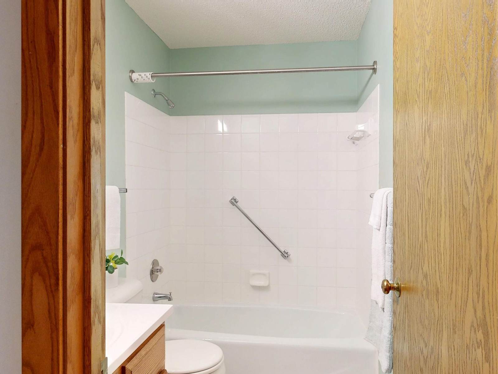 Older hall bathroom with a white tile tub and shower combination, a diagonal grab bar and an oak door, a typical hall tub before a remodel