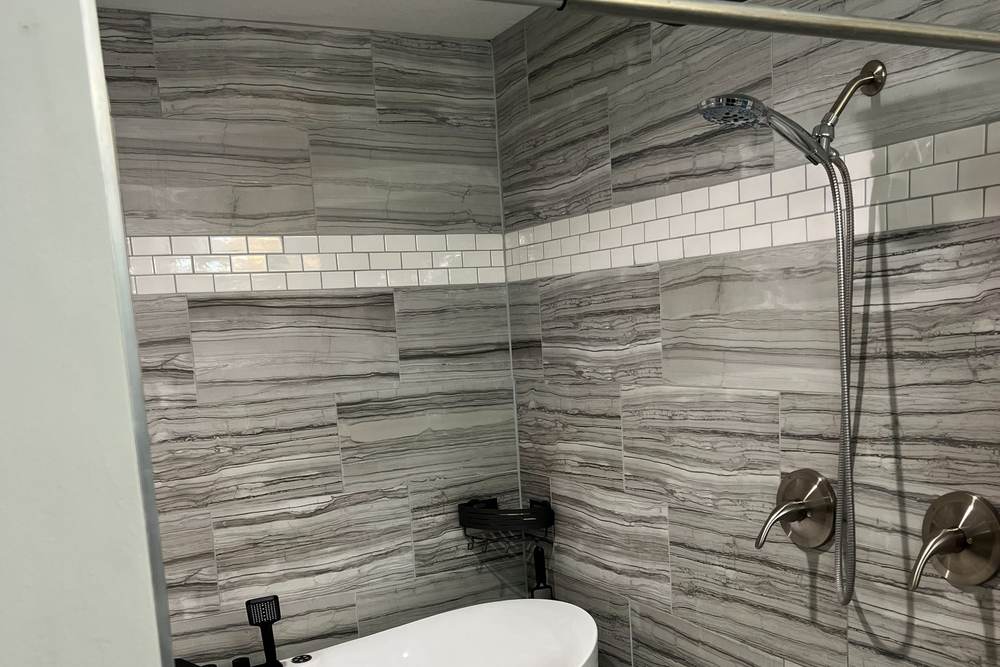 Overhead rain head, a hand shower and two shower handles on the tiled wall beside a freestanding tub, in the south Tulsa wet room we built