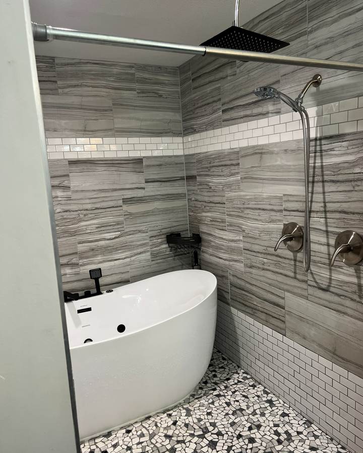 A freestanding tub beside a handheld shower on gray plank tile with a subway tile band