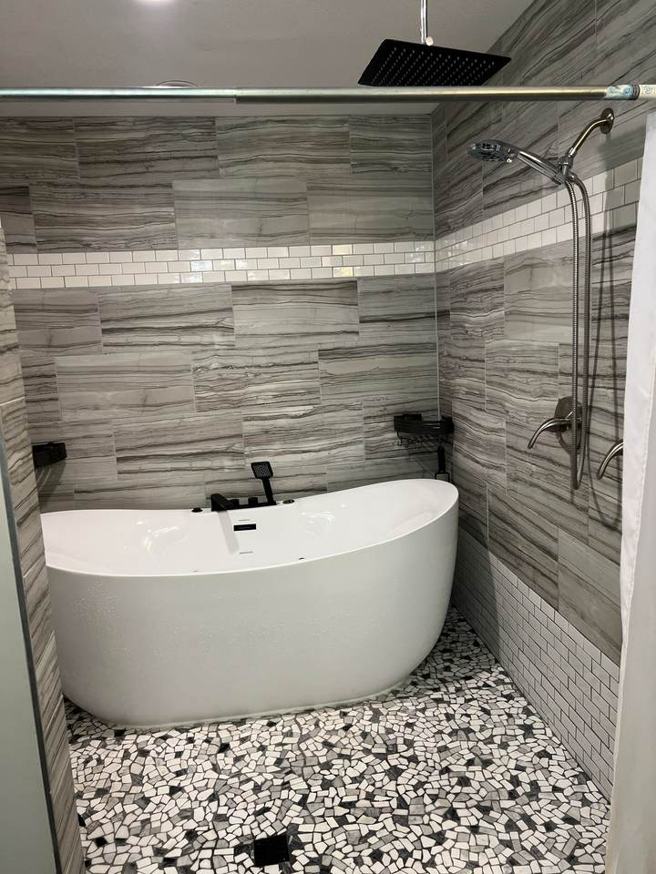 Freestanding soaking tub in a tiled wet room with a pebble floor and a white subway tile band, in a south Tulsa home
