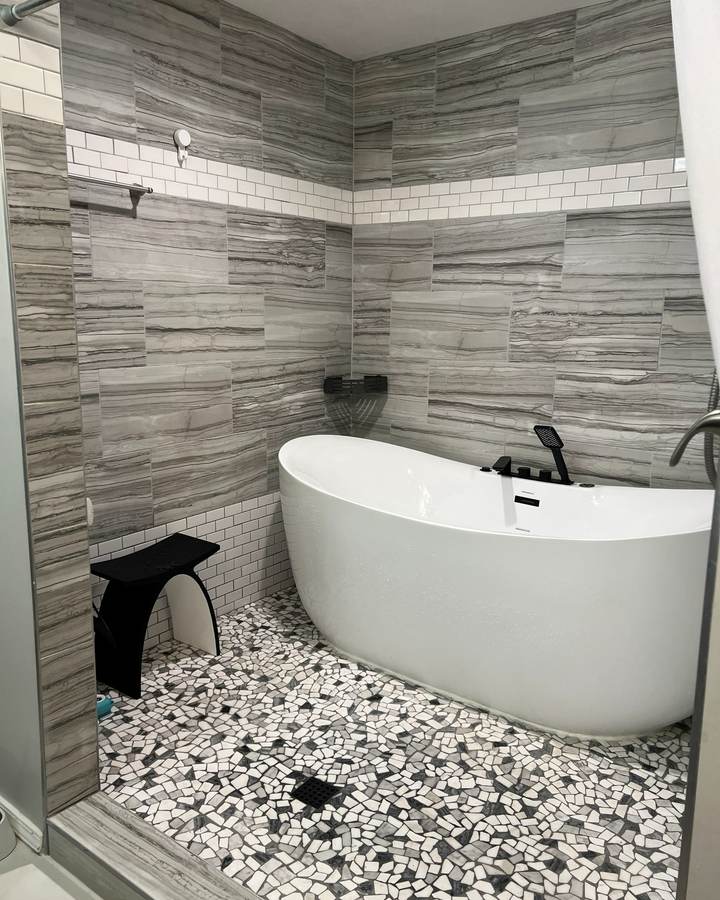 A white soaking tub with black fixtures and a black stool on a pebble mosaic floor