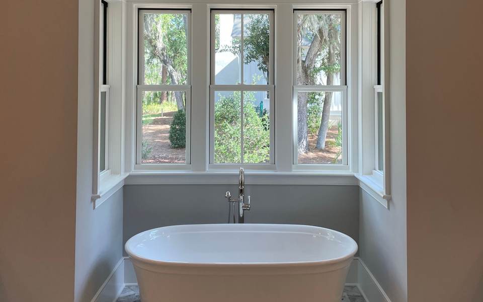 Freestanding soaking tub in a bay window that looks out on trees, over a marble-look herringbone floor