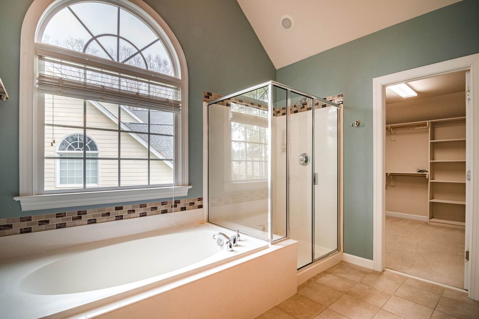 Drop-in garden tub under an arched window beside a framed glass shower stall, with a walk-in closet through the door