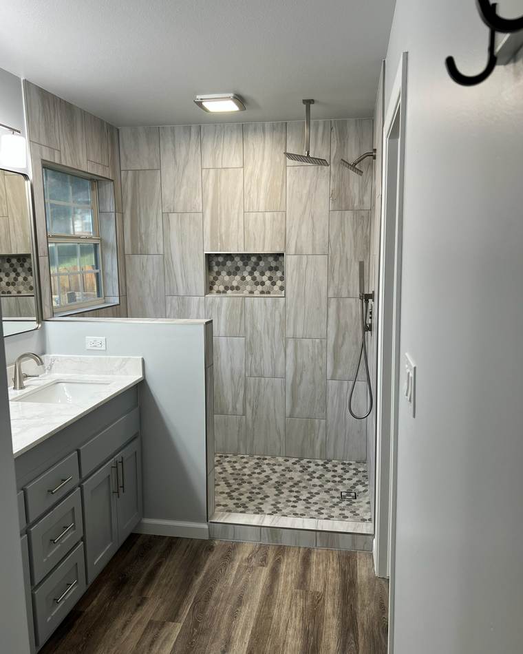Tiled walk-in shower behind a half wall beside a gray double vanity, in a master bathroom we remodeled in east Tulsa