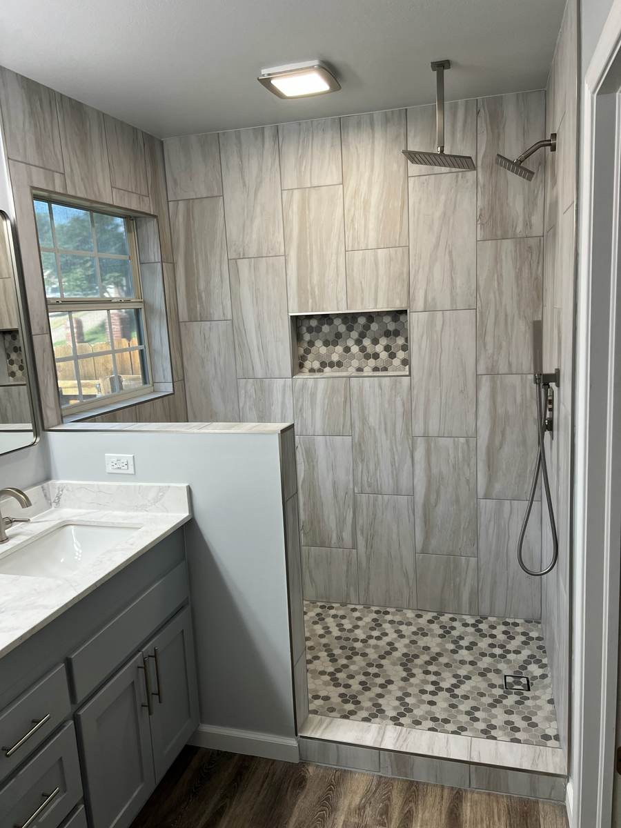 After: a tiled walk-in shower with a rain head, a hex-tile niche and a half wall beside a gray vanity, in the same Tulsa master bathroom