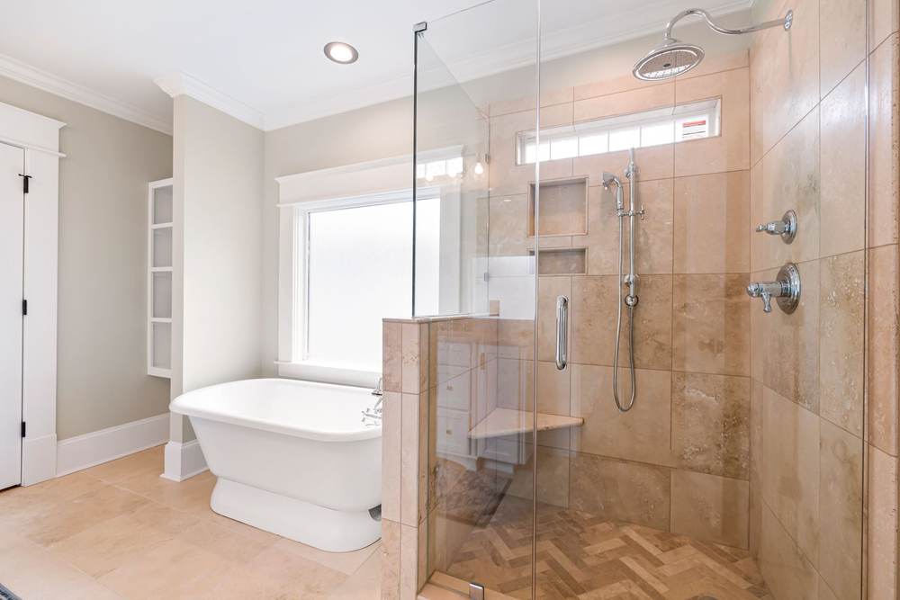 Glass walk-in shower with a tiled half wall, a corner bench and a rain head beside a pedestal freestanding tub