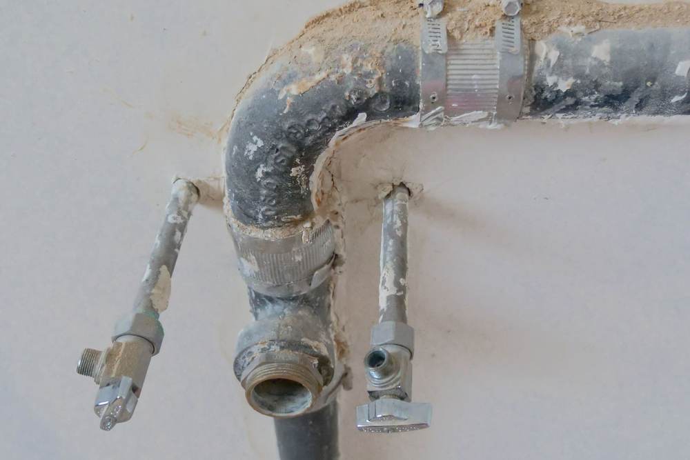 Old metal drain pipe and worn supply lines with shutoff valves coming out of an opened bathroom wall, the kind of plumbing replaced during a remodel