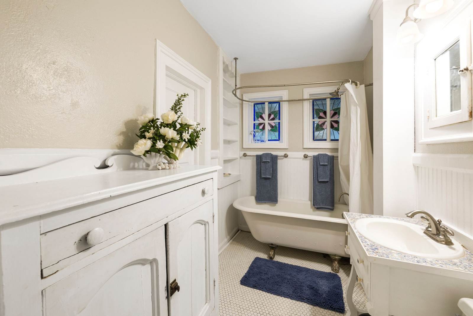 Older bathroom with a clawfoot tub under two stained glass windows, a white hex tile floor and painted wainscot, the kind of room many prewar houses still have