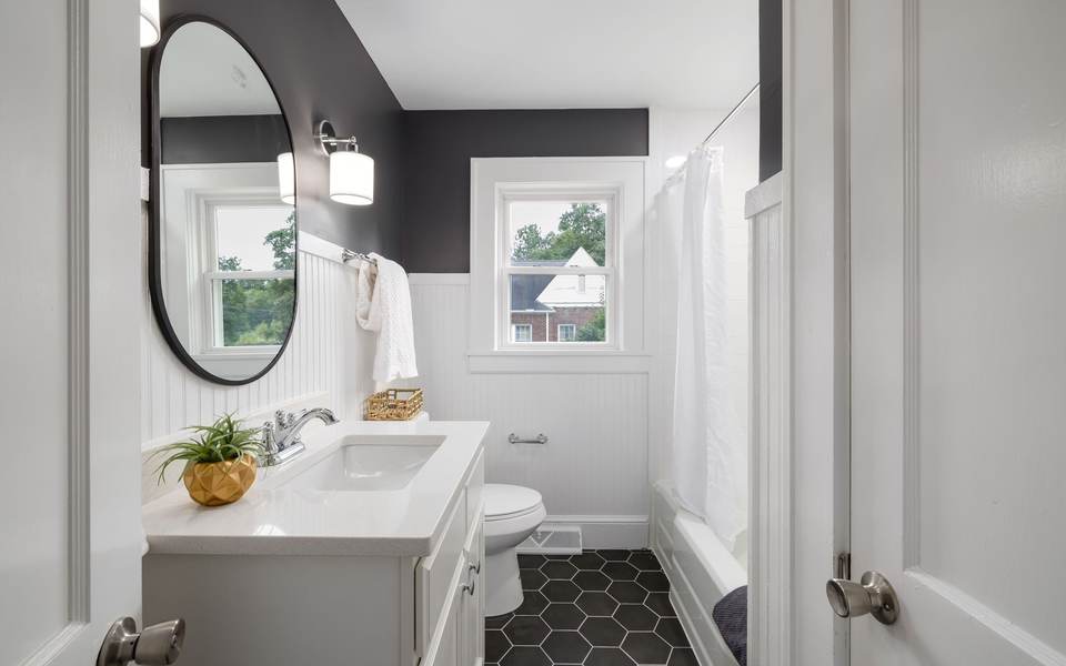 Bathroom with white beadboard walls, a white vanity under an oval mirror, a tub behind a white curtain and a black hex tile floor