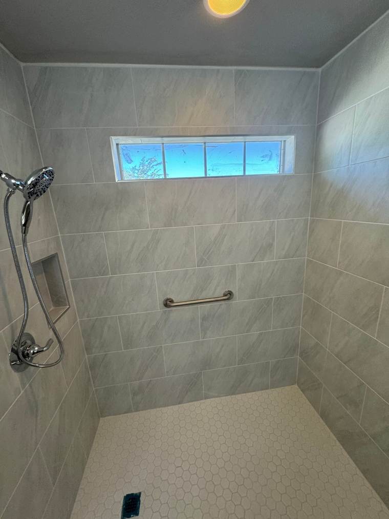 Roll-in shower with large gray tile, a white hex floor, a niche and a transom window where a corner jetted tub used to be