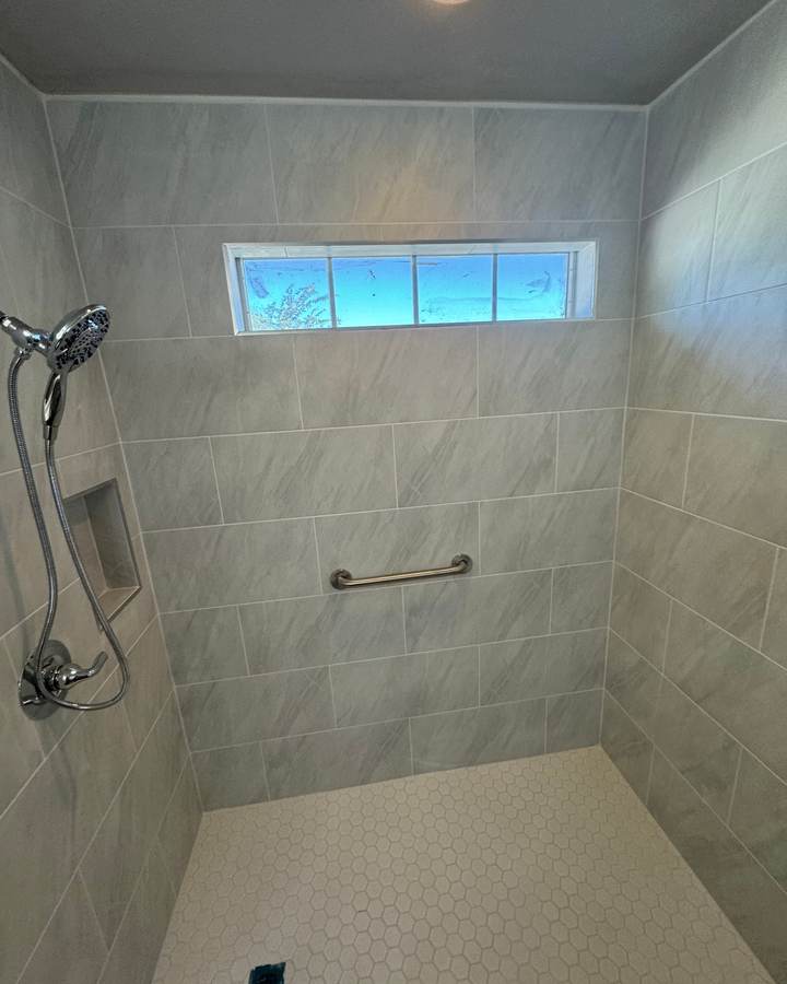 A roll-in shower with large gray tile, a white hex floor, a niche and a transom window