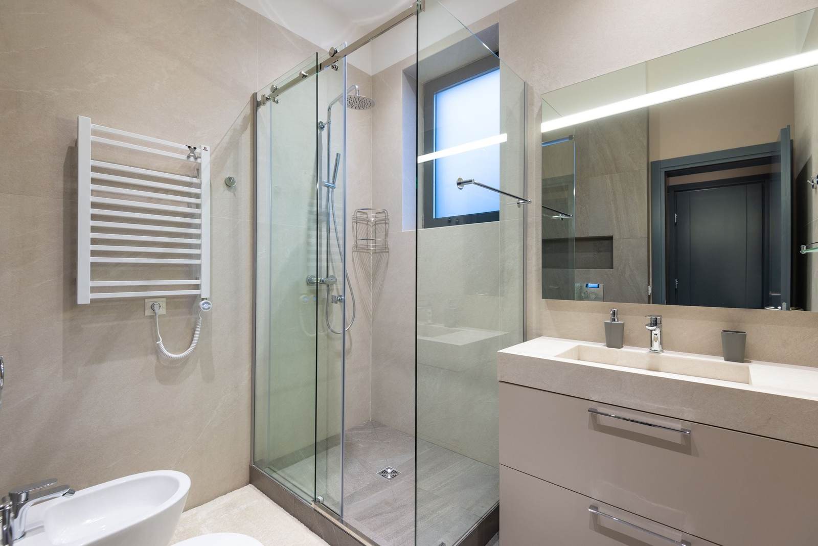 Glass shower with sliding doors, a rain head and a hand shower over a low tiled floor, beside a beige vanity with a lit mirror
