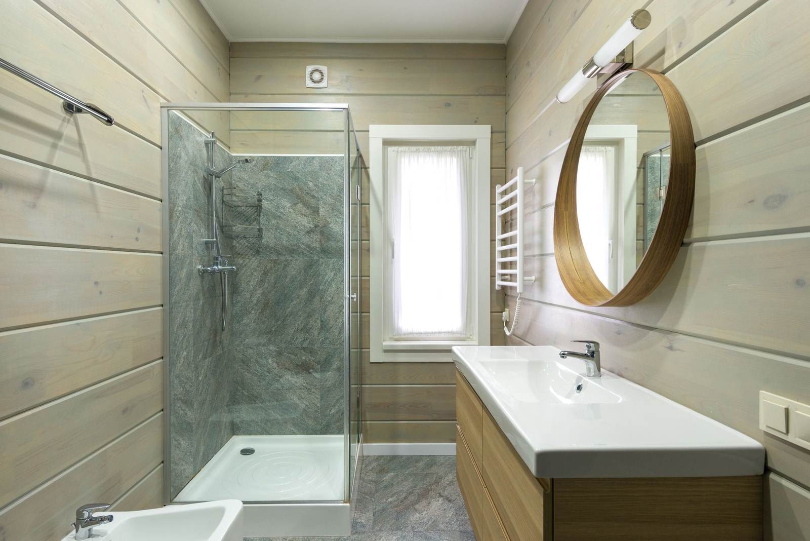 Glass corner shower with stone-look wall panels beside a small window, in a bathroom with painted wood plank walls and a round wood mirror