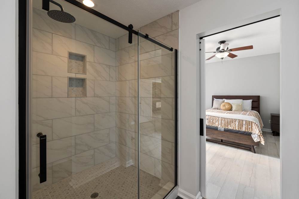 Sliding glass shower door on a black roller bar, in front of a tiled shower with two niches