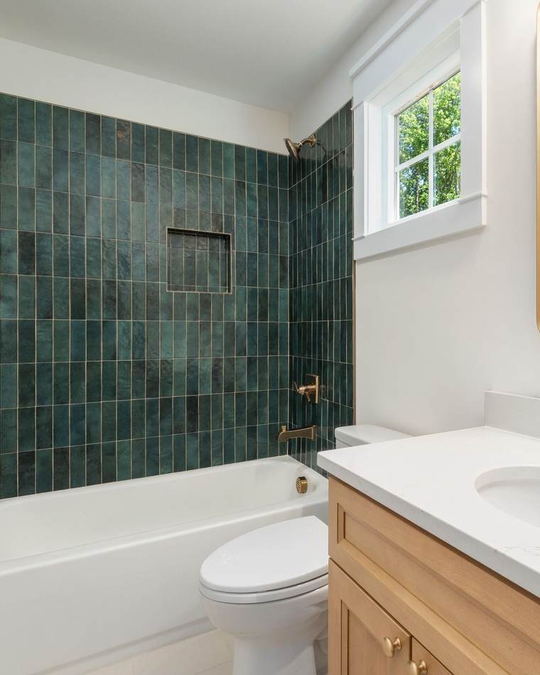 Small bathroom with green glazed tile around a tub and shower, a tiled niche, a window and a wood vanity with a quartz top