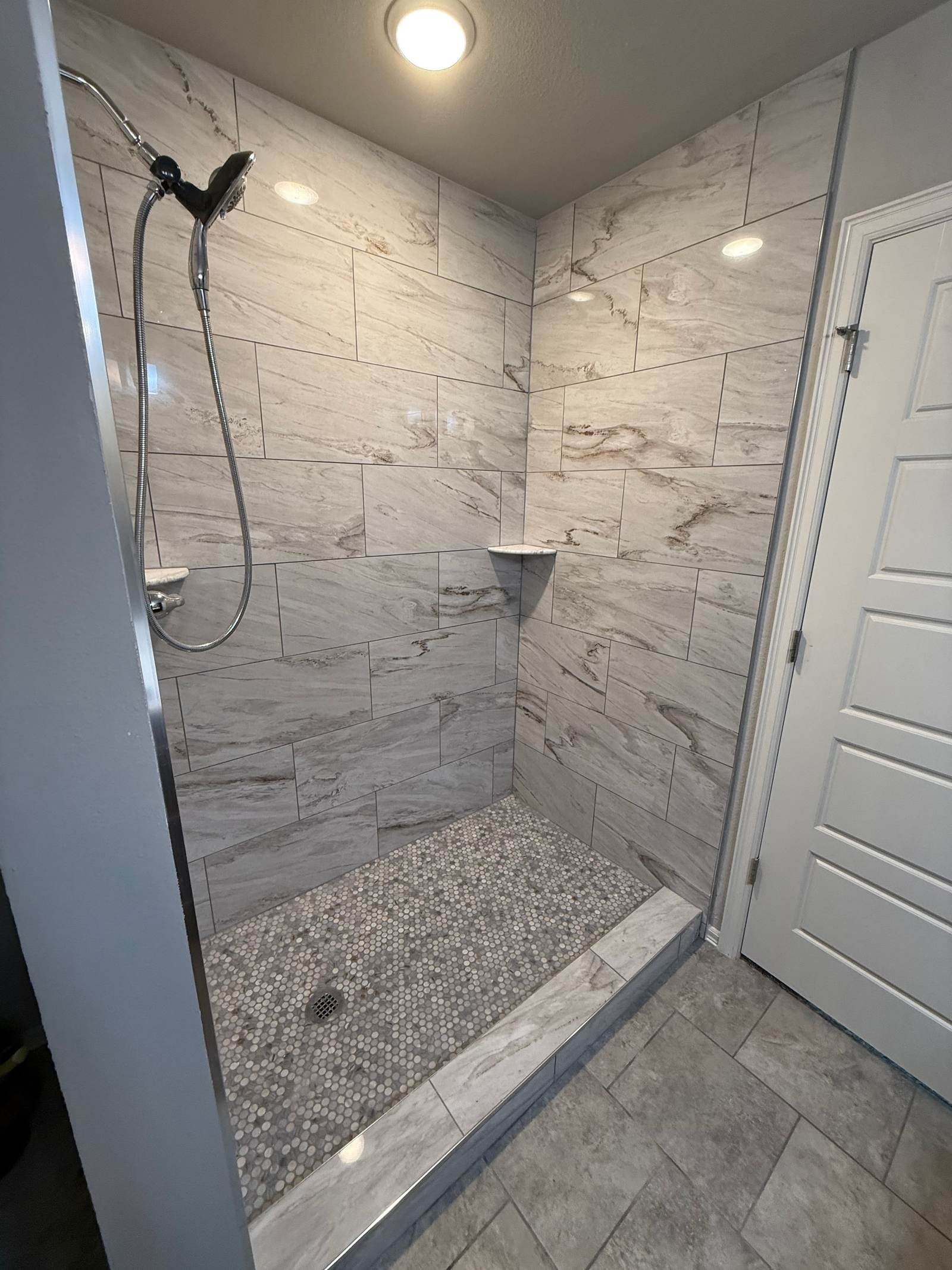 Tiled walk-in shower with marble-look walls, corner shelves and a pebble floor, built where a fiberglass tub and shower unit used to be, in a Bixby home we remodeled