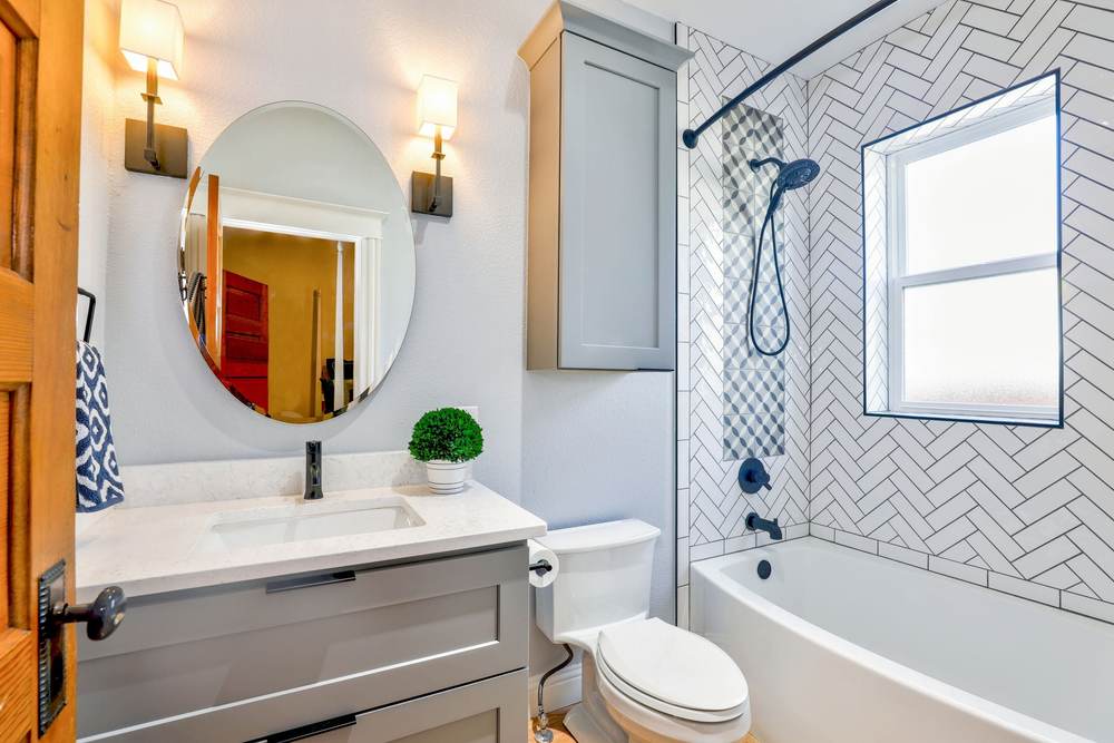 Small bathroom with a tub and shower in white herringbone tile under a window, beside a gray vanity and an oval mirror