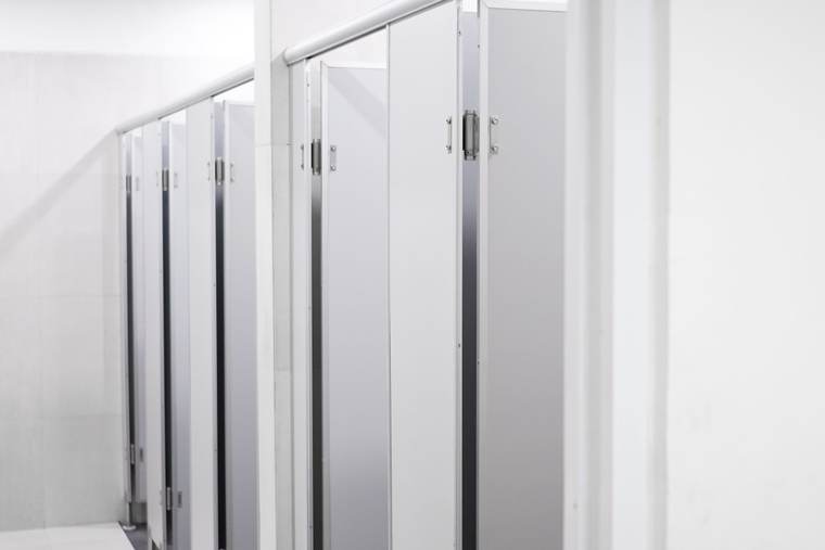 Row of white laminate toilet partitions with stainless hardware in a bright commercial restroom