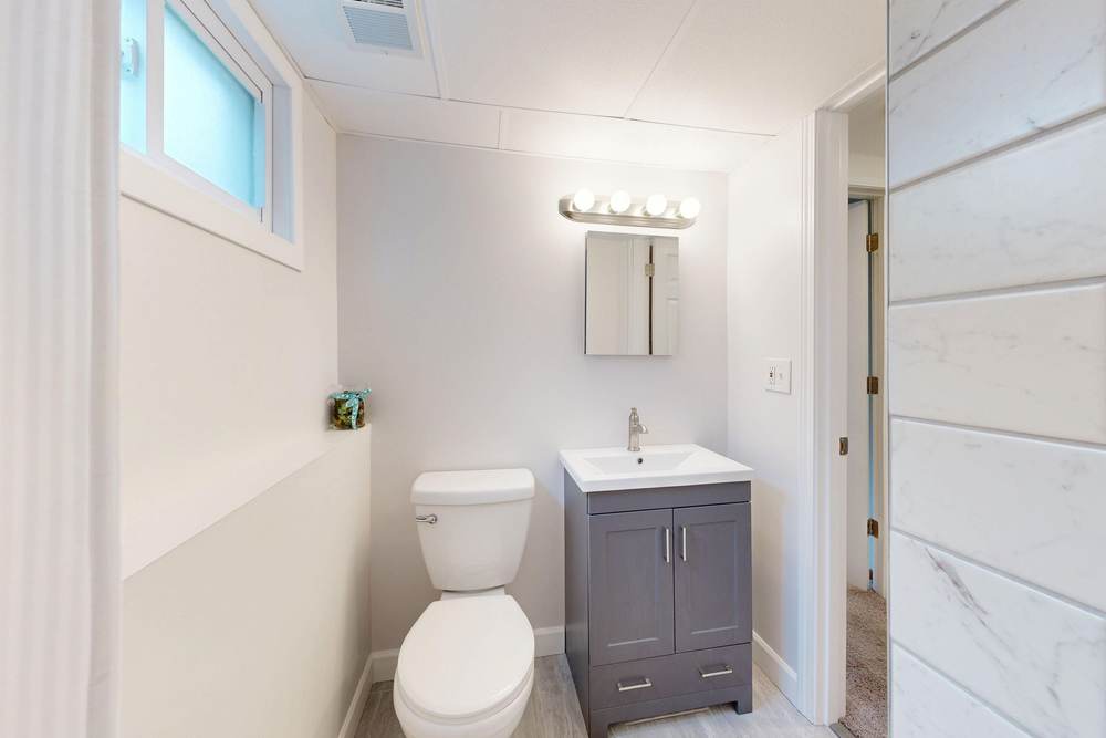 White two-piece toilet beside a small gray vanity in a compact bathroom with a high window