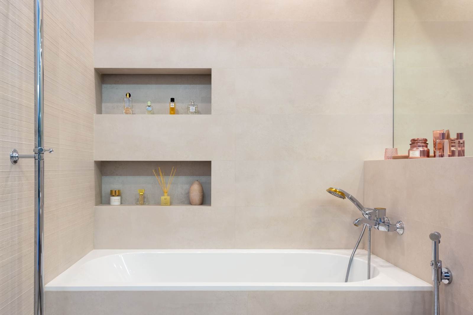 Tiled tub and shower with two recessed niches, a wall-mounted tub faucet with a hand shower, and a glass screen