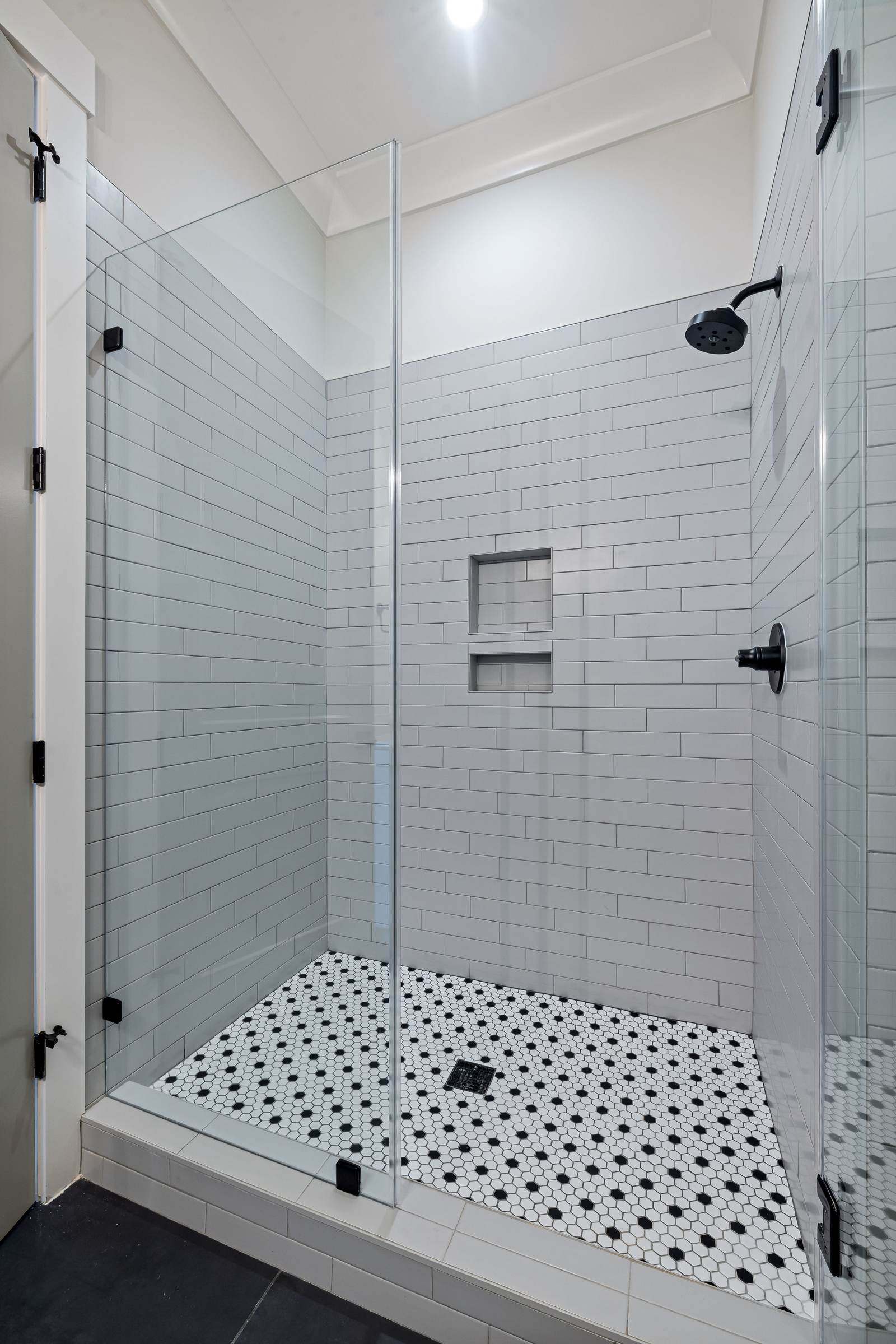 Compact tiled walk-in shower with two stacked niches, white subway tile, a black and white mosaic floor and frameless glass