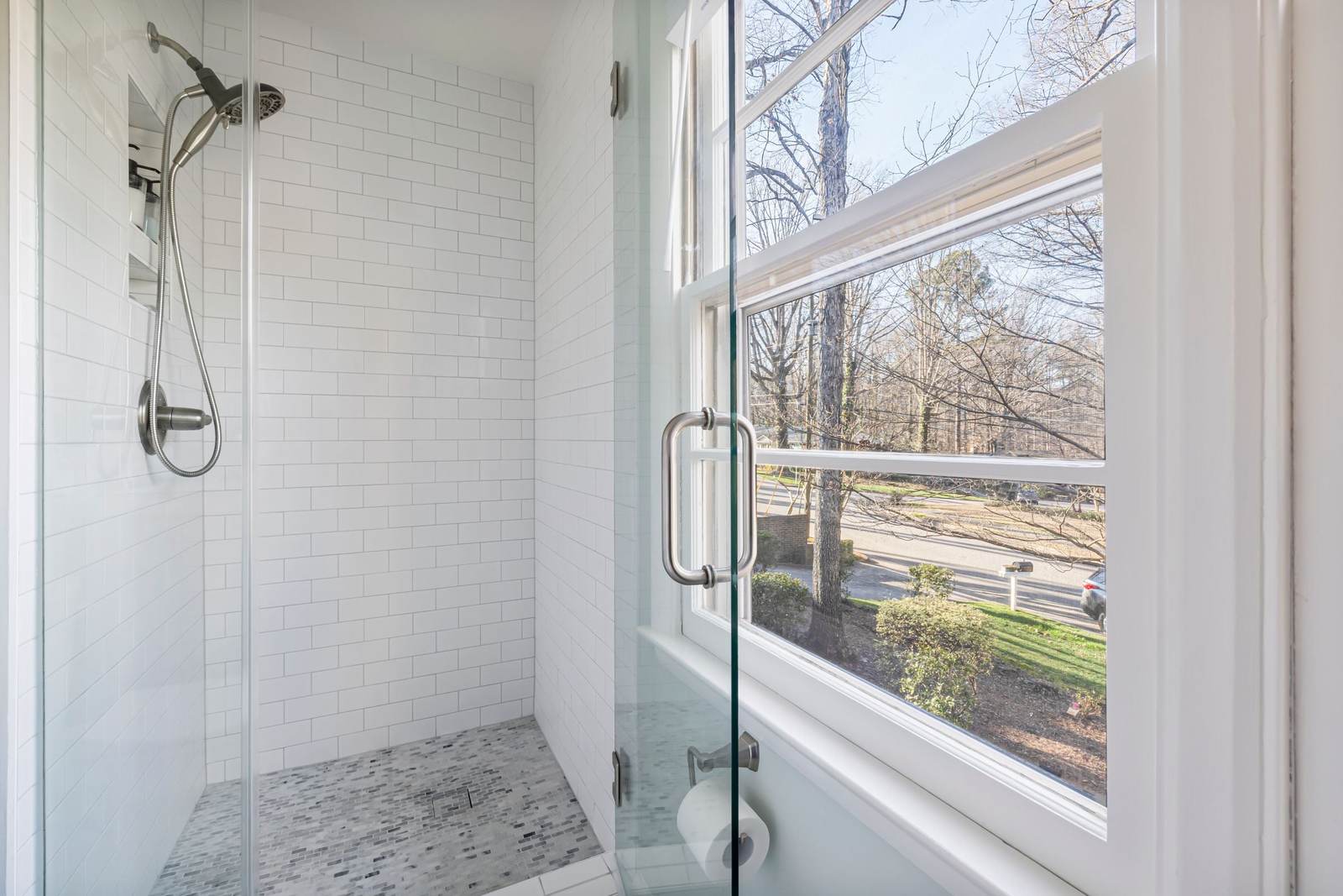 Frameless glass door on a white subway tile shower, beside a window looking out on bare trees