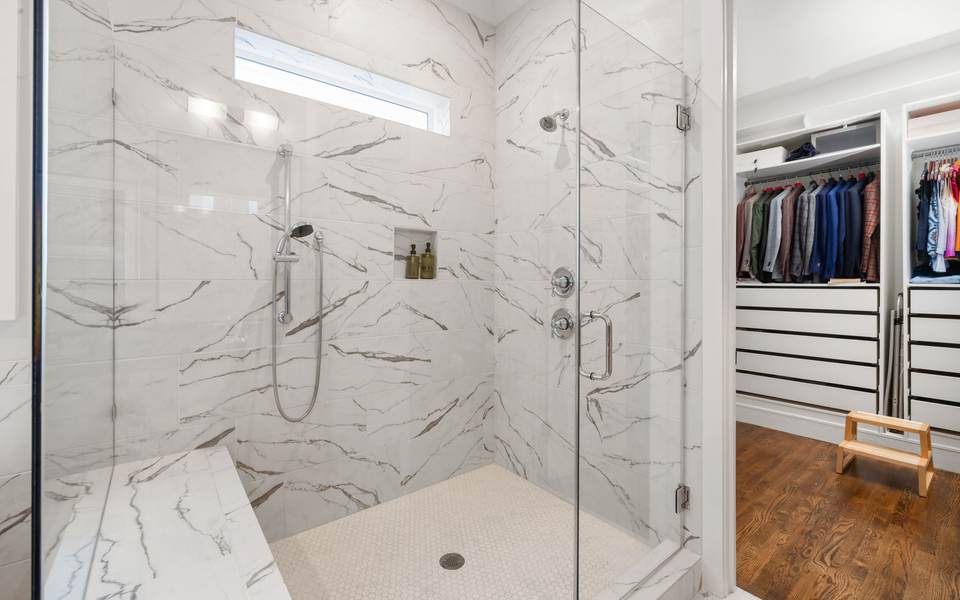 Marble-look tiled walk-in shower with a built-in bench, a hand shower and a frameless glass panel beside a walk-in closet