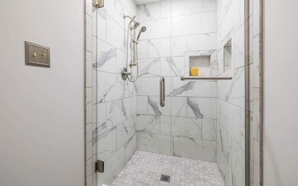 Marble-look tiled walk-in shower with a grab bar, a hand shower on a sliding rail, a recessed niche and a mosaic floor