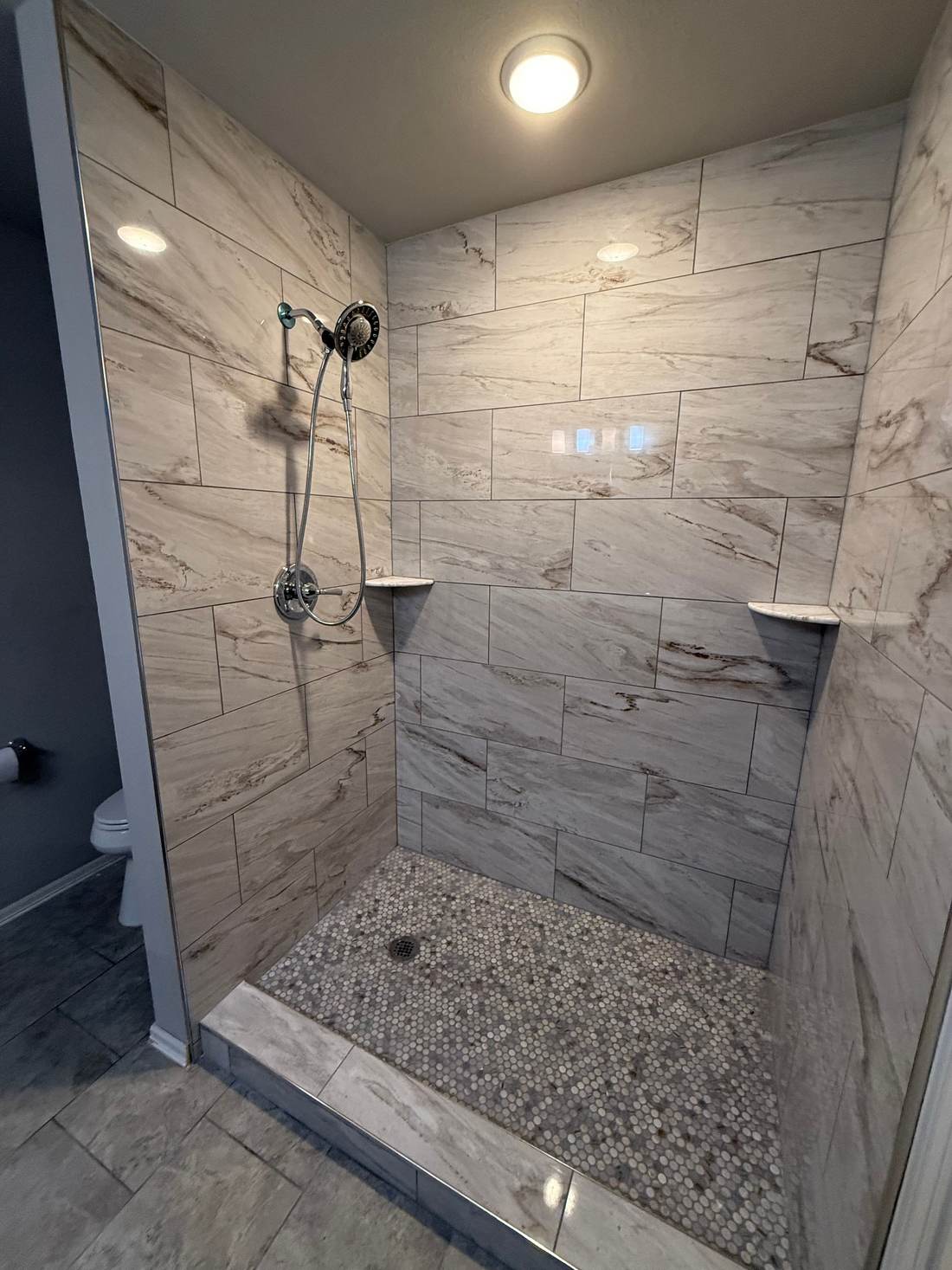 Walk-in shower with a low tile curb, marble-look walls and a pebble mosaic floor where a tub and shower combo used to be