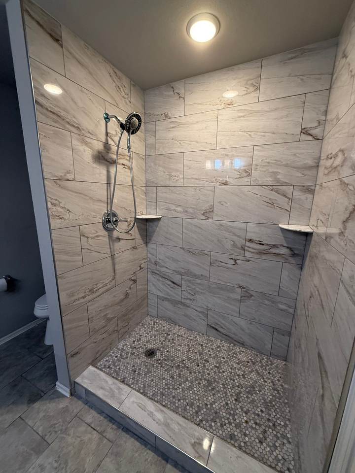 A walk-in shower with a low tile curb, marble-look walls and a pebble mosaic floor