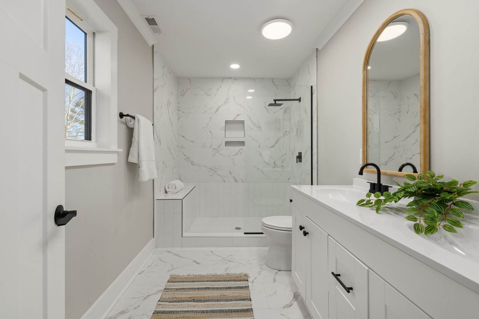 Walk-in shower with marble-look tile, a tiled bench, a niche and a glass panel, beside a white vanity and an arched wood mirror