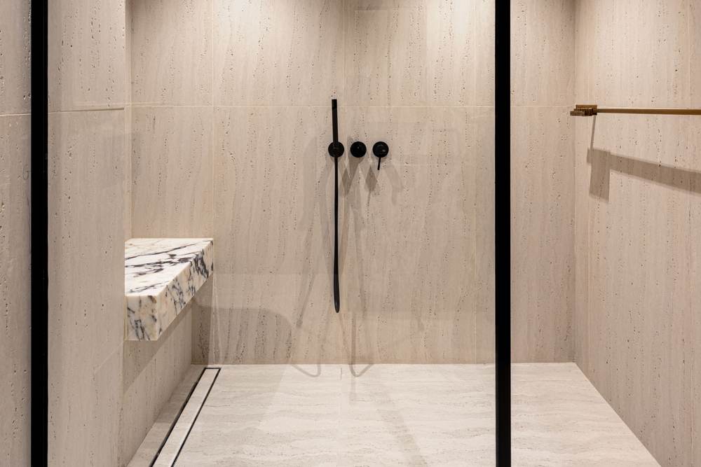 Curbless walk-in shower with a linear drain, matte wood-look porcelain tile and black fixtures behind a black-framed glass panel