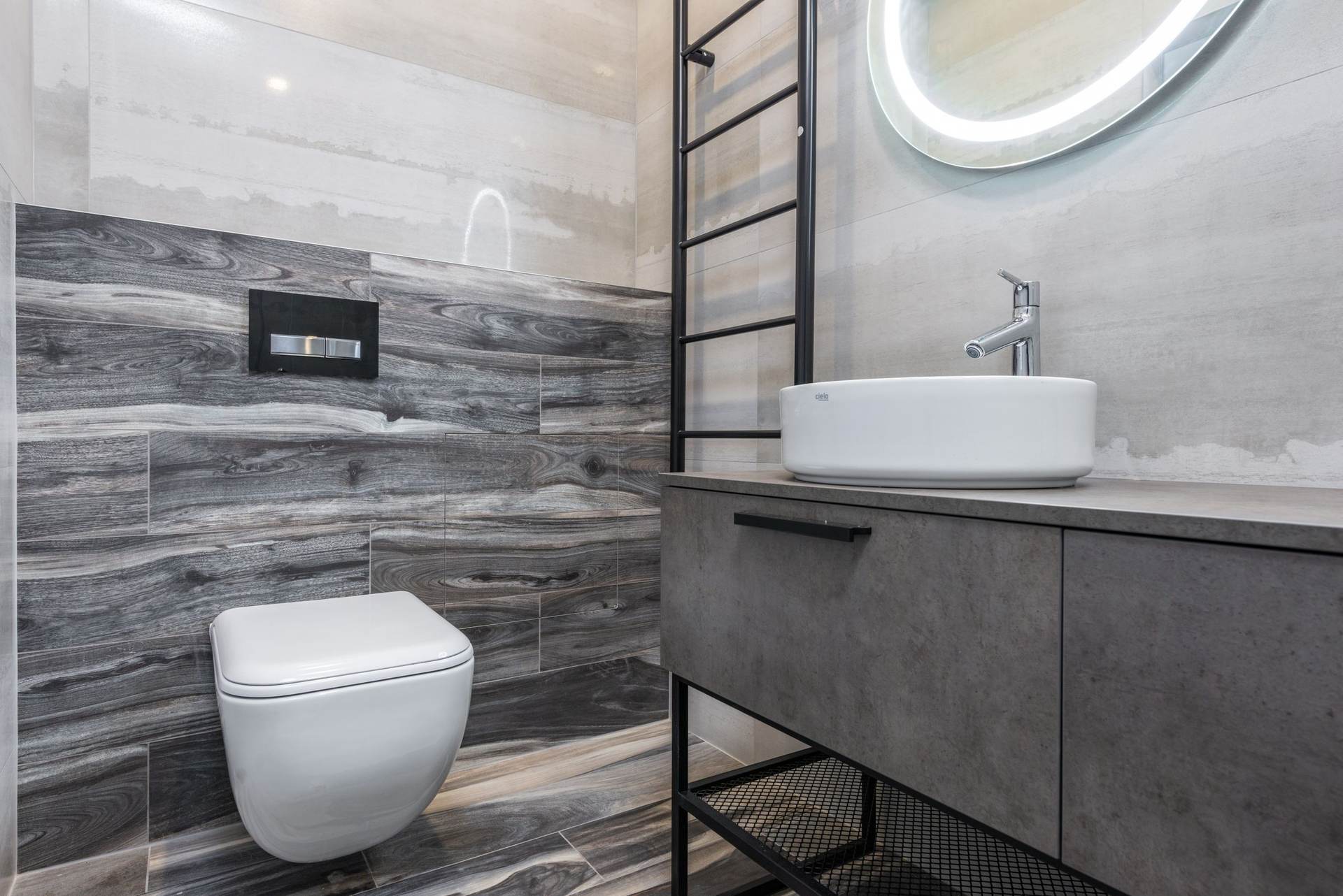 Wall-hung toilet with an in-wall flush plate on gray wood-look tile beside a floating vanity