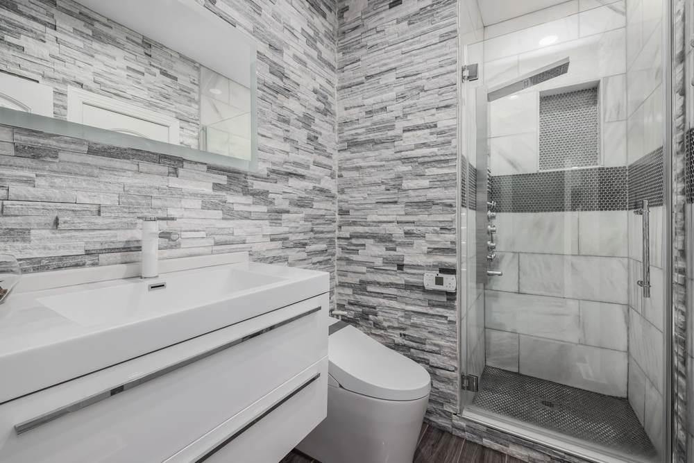 Toilet with an electronic bidet seat beside a white floating vanity, stacked stone tile walls and a glass shower with a penny tile floor
