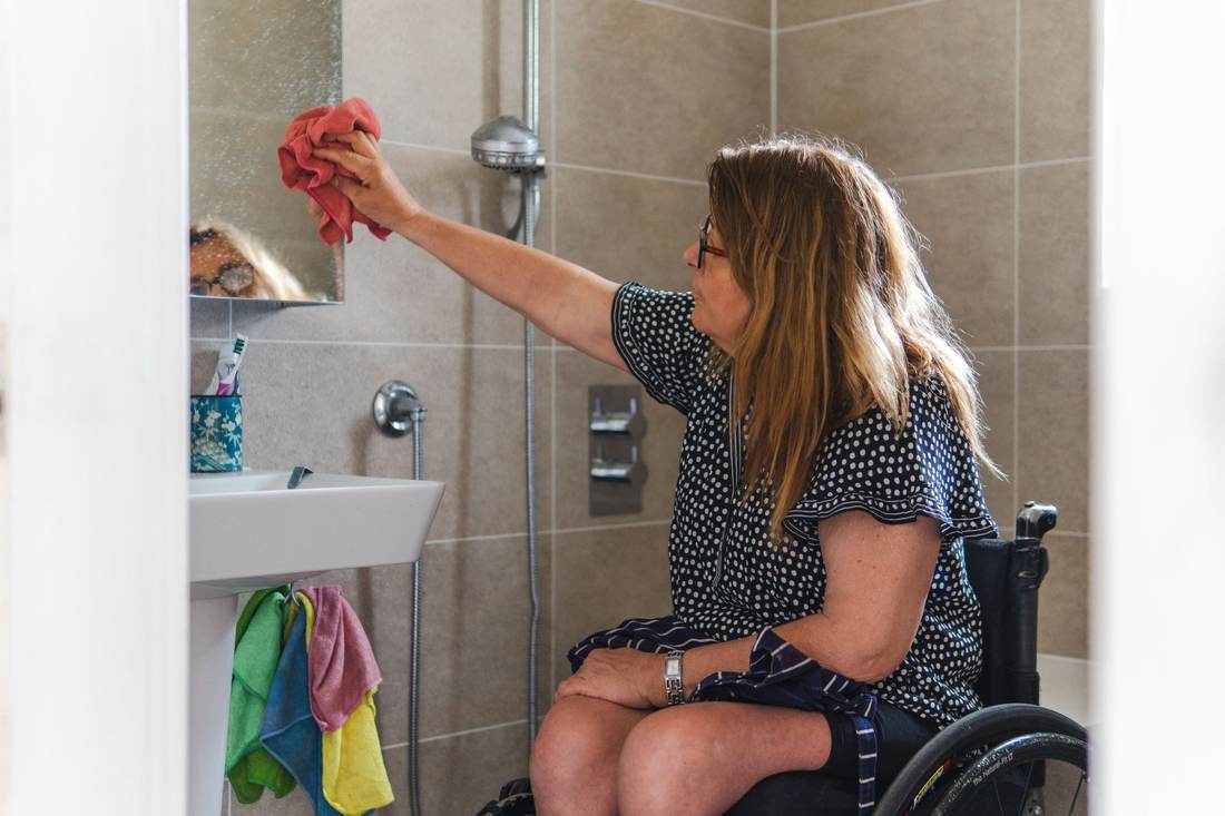 Woman in a wheelchair wiping the mirror in an accessible bathroom with a shower valve on the wall beside her