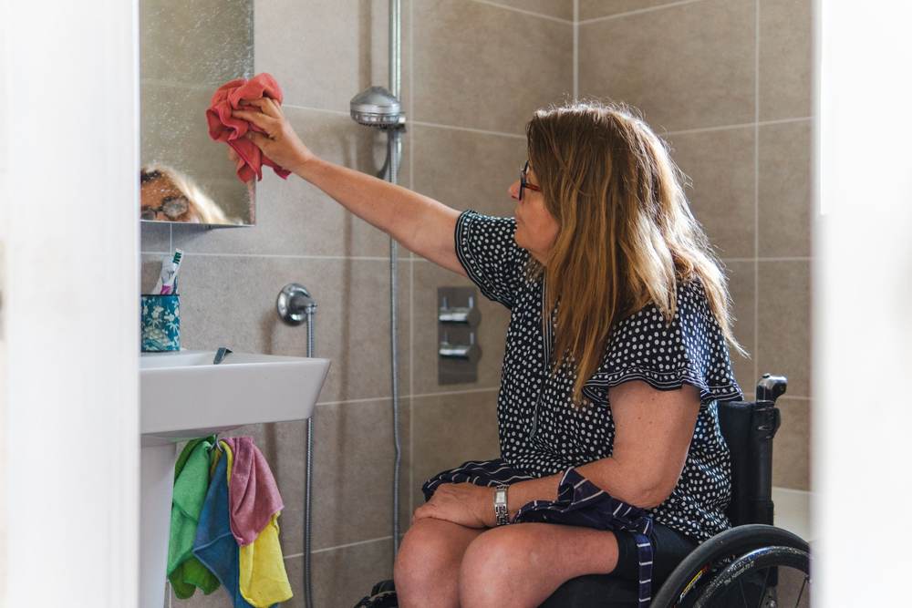 Woman in a wheelchair wiping a bathroom mirror above a wall-hung sink, beside a shower with a hand shower